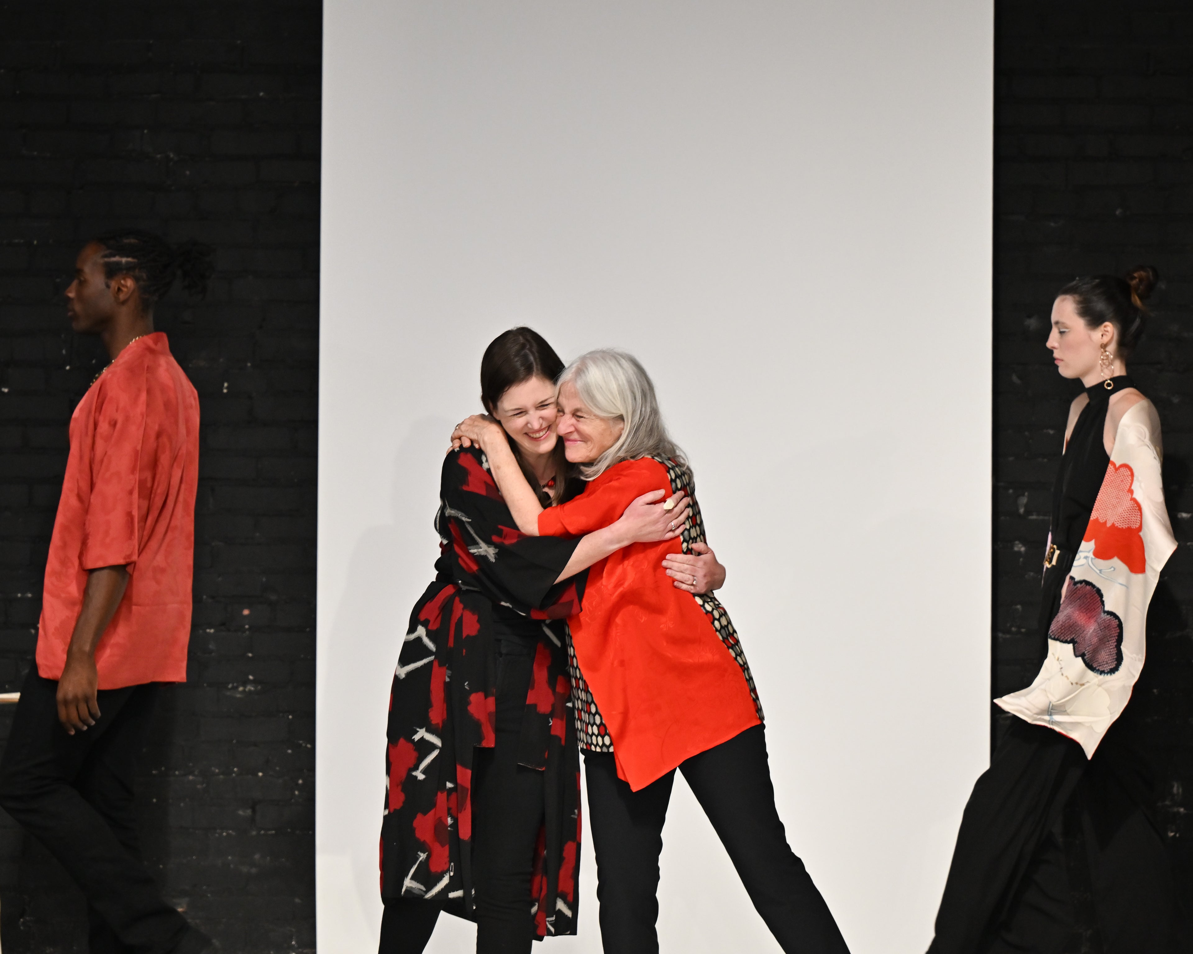 Two women hugging on a white background with fashion models in the foreground.