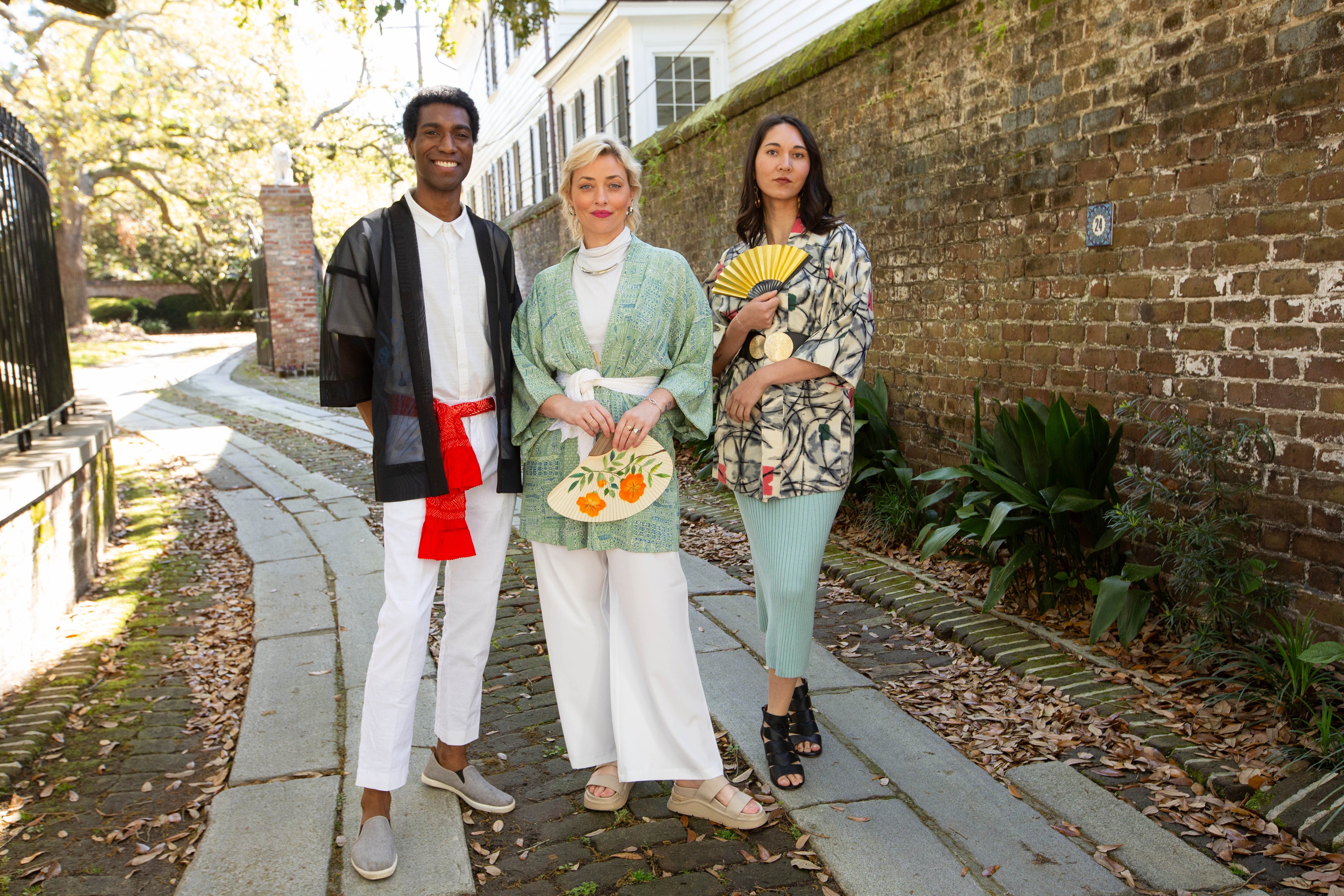 Three people standing on a sidewalk with a brick wall and greenery in the background