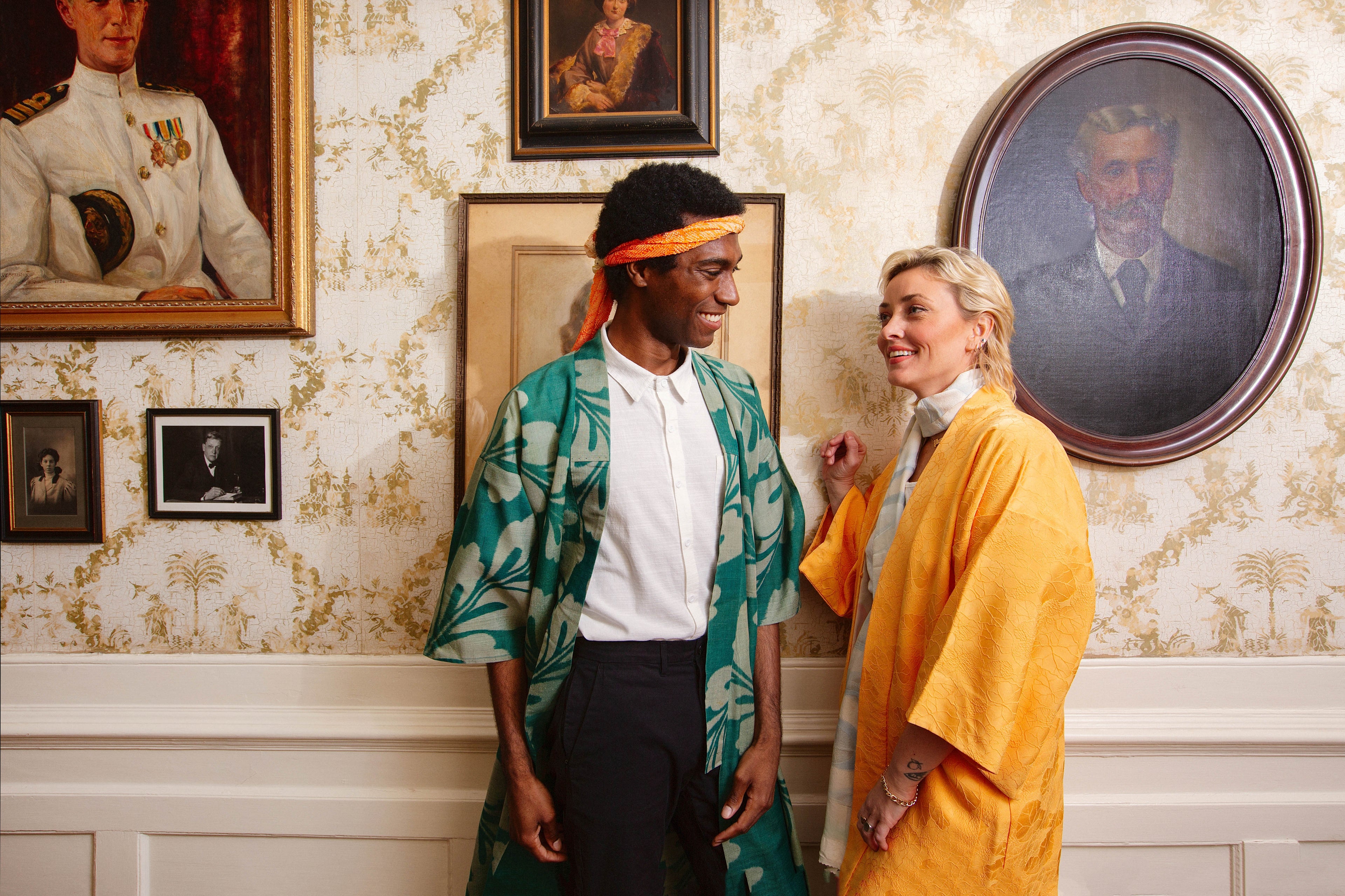 Two people standing in a room with ornate wallpaper and framed portraits.