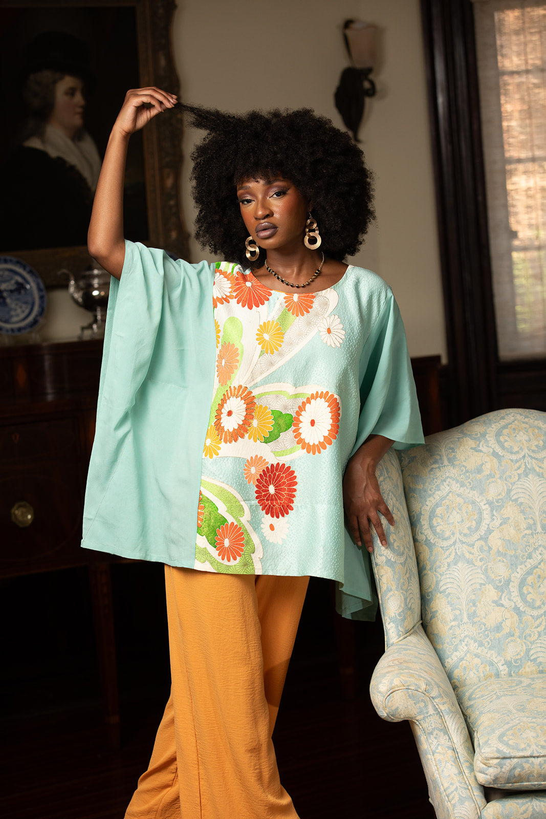 Woman in a floral top and orange pants standing in a room with a chair and mirror.